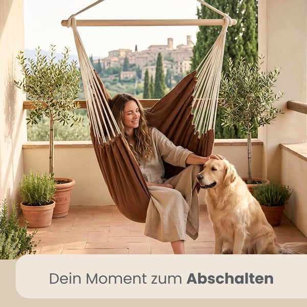 Frau sitzt in braunem Hängesessel aus Stoff auf hellem Balkon und streichelt einen Hund. Im Hintergrund sind Topfpflanzen und Stadtblick zu sehen.