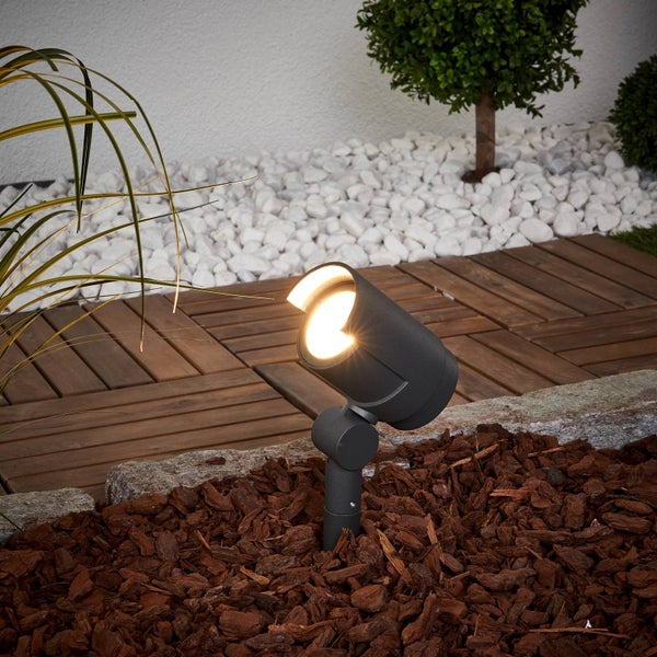 Anthrazitfarbener LED-Gartenstrahler mit Erdspieß in einem Beet mit Rindenmulch neben einer Holzterrasse.