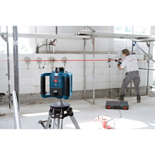 Bosch Logo. Der Linienlaser projiziert eine rote Linie an eine Mauer, während eine Person mit einer Bohrmaschine arbeitet.