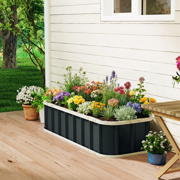 Rechteckiges anthrazitfarbenes Hochbeet aus Metall mit Blumen bepflanzt auf einer Holzterrasse neben einer Hauswand in einem hellen Garten.
