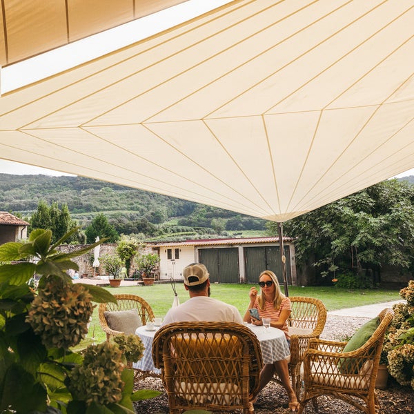 Großes beiges Sonnensegel über einer Sitzgruppe aus Rattan mit zwei Personen im hellen Garten mit Blick auf grüne Hügel.