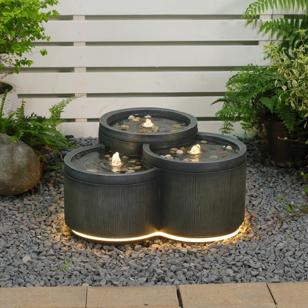 Dreistufiger Gartenbrunnen mit beleuchteten Wasserspielen und Sockelbeleuchtung auf Kies vor einem Holzzaun und Pflanzen.