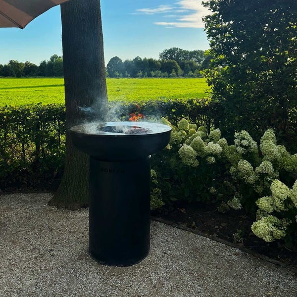 Schwarzer zylindrischer Außengrill von BonFeu mit runder Grillplatte, auf der Fleisch über offenem Feuer in einem Garten gegart wird.
