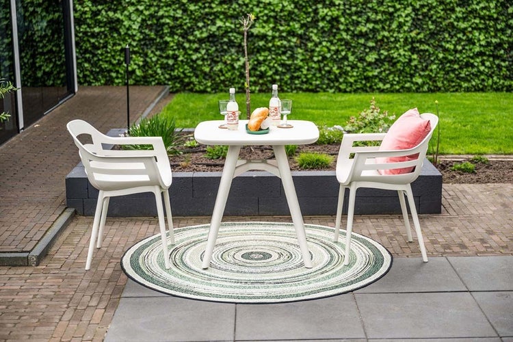 Gartenmöbel-Set mit weißem Tisch und zwei Stühlen auf einem runden Außenteppich in einem hellen, gepflegten Garten.