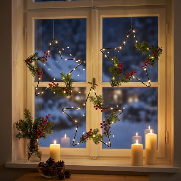 Drei beleuchtete Weihnachtssterne zum Aufhängen im Fenster, verziert mit Tannenzweigen und Beeren, dazu Kerzen und Tannenzapfen auf dem Fensterbrett.