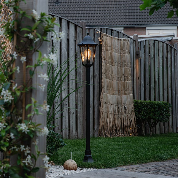 Schwarze Wegeleuchte im klassischen Laternen-Stil auf einer Rasenfläche vor einem Holzzaun im Garten.