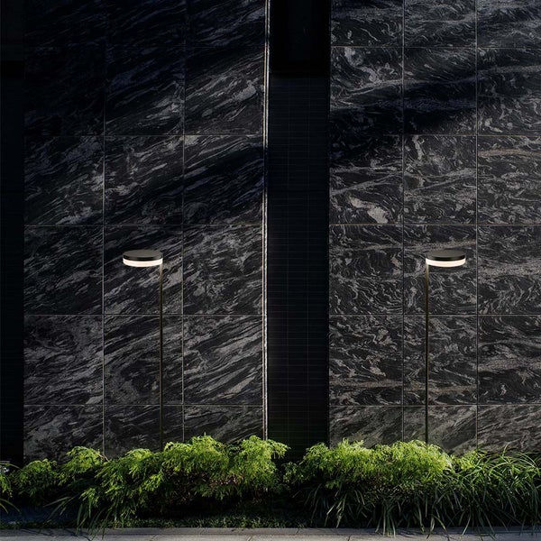 Zwei Außenleuchten beleuchten eine dunkle Fassade und grüne Pflanzen.