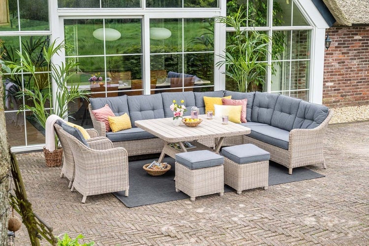 Lounge-Set aus Polyrattan mit Ecksofa, Sesseln, Hockern und Tisch auf einer Terrasse vor einem Haus mit großen Fenstern.