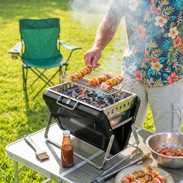 Tragbarer Holzkohlegrill auf einem Tisch im Garten mit Fleischspießen, Grillzange und Campingstuhl im Hintergrund.
