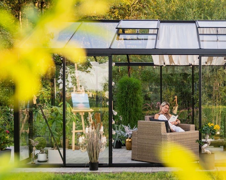 Modernes Gewächshaus mit schwarzem Rahmen, eingerichtet als heller Gartenraum mit Korbsessel, Pflanzen und einer Staffelei.