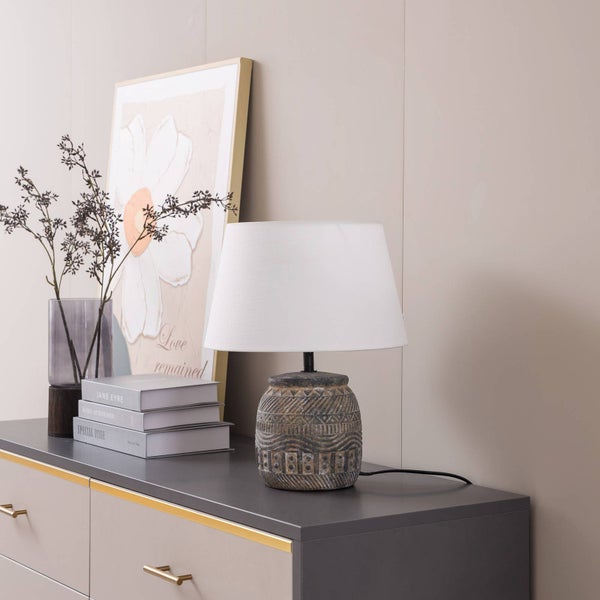 Tischleuchte mit gemustertem Keramikfuß und weißem Stoffschirm auf einer Kommode neben einer Vase und Büchern.