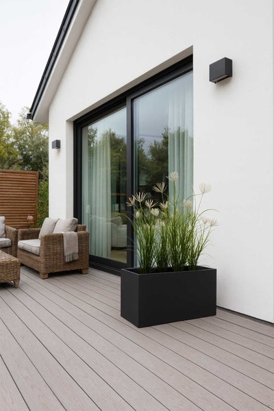 Moderner Terrassenbereich mit schwarzem Pflanzkübel, Ziergräsern, hellen Terrassendielen und Rattan-Gartenmöbeln vor einer weißen Hauswand.