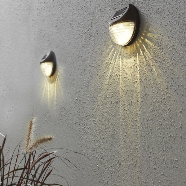 Zwei Solarwandleuchten an einer Wand mit Pflanzen im Vordergrund