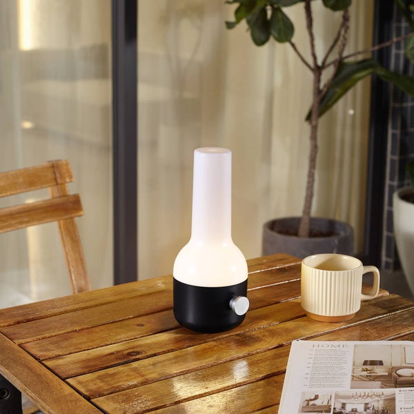 Tragbare Tischleuchte in Schwarz und Weiß im Flaschendesign auf einem Holztisch mit Tasse und Zeitschrift in heller, organisierter Umgebung.