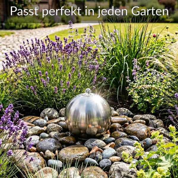 Edelstahl-Gartenbrunnen Kugel auf Steinen, umgeben von Lavendel und Gräsern in einem Gartenbeet.
