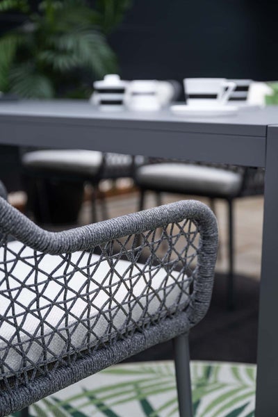 Detailansicht eines modernen Gartenstuhls mit dunkelgrauem Seilgeflecht und hellem Sitzkissen an einem grauen Esstisch.