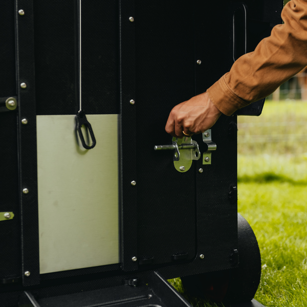 Eine Hand bedient einen Metallriegel an der Tür eines schwarzen Hühnerstalls.
