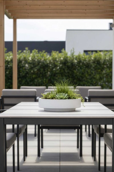 Weiße runde Pflanzschale mit Sukkulenten und Gras auf einem modernen Gartentisch unter einer Holzpergola auf einer hellen Terrasse.