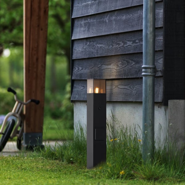 Schwarze Außenstehleuchte in Säulenform mit Filament-Leuchtmittel vor einer Holzfassade im Garten.