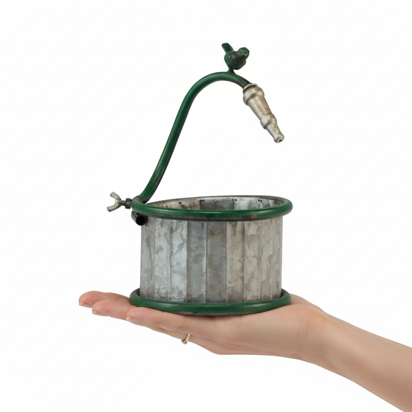 Kleiner Übertopf aus verzinktem Metall mit grünem, geschwungenem Griff und kleiner Vogelfigur, in einer Hand gehalten.