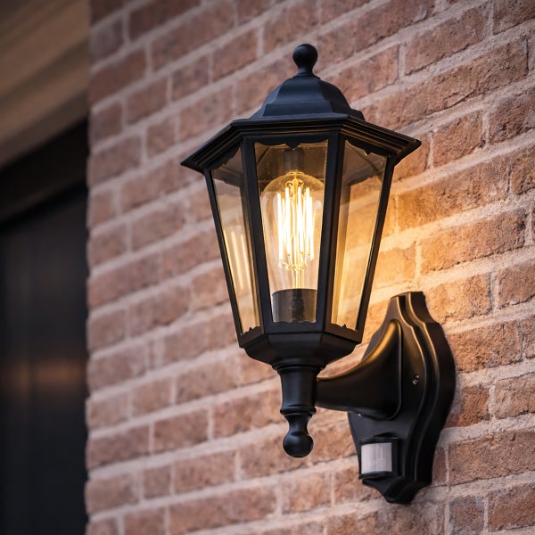 Klassische schwarze Außenwandleuchte mit Bewegungsmelder und leuchtender LED-Filament-Lampe an einer Backsteinwand.