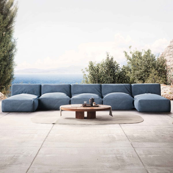 Modernes blaues Modulsofa auf einer hellen Steinterrasse mit einem runden Couchtisch aus Holz, hellem Teppich und weitem Meerblick.