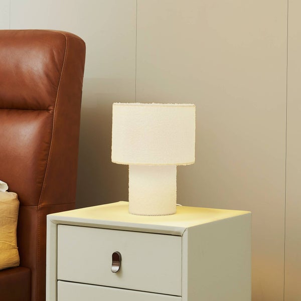 Eingeschaltete Tischleuchte mit strukturiertem Bouclé-Schirm und Standfuß auf einem weißen Nachttisch neben einem braunen Lederbett.