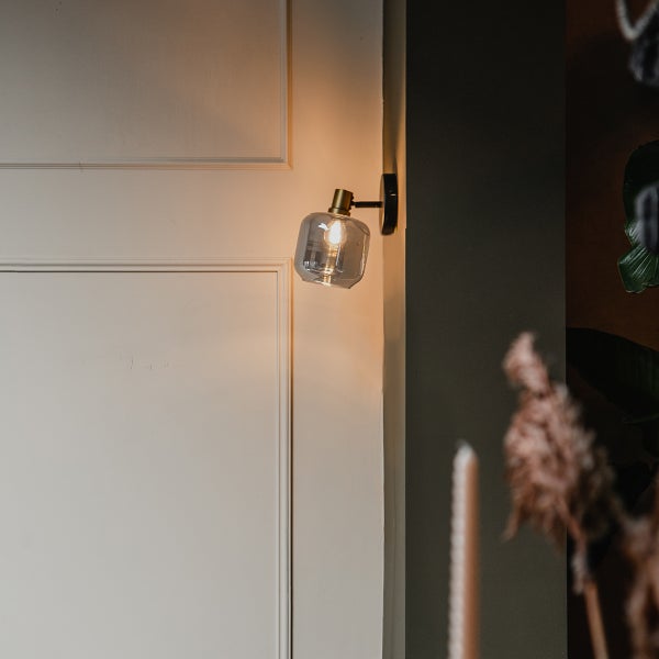 Moderne Wandleuchte mit grauem Glasschirm und Messingarm an einer Paneelwand.