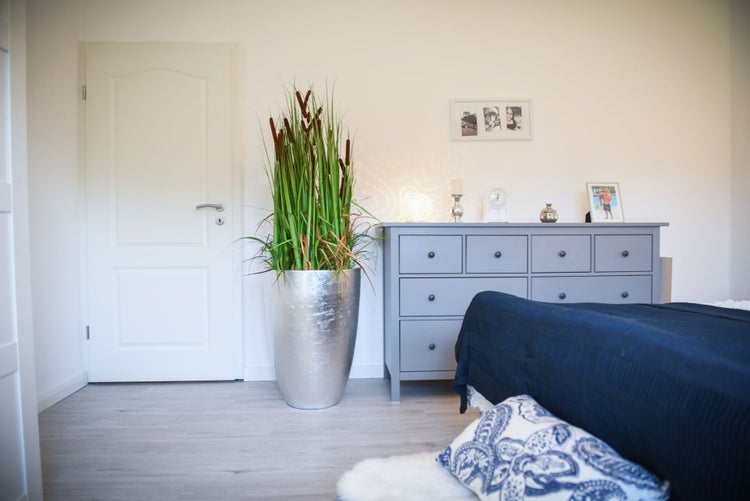Moderner silberner Pflanzkübel mit künstlichem Rohrkolben in einem hellen Schlafzimmer neben einer grauen Kommode.