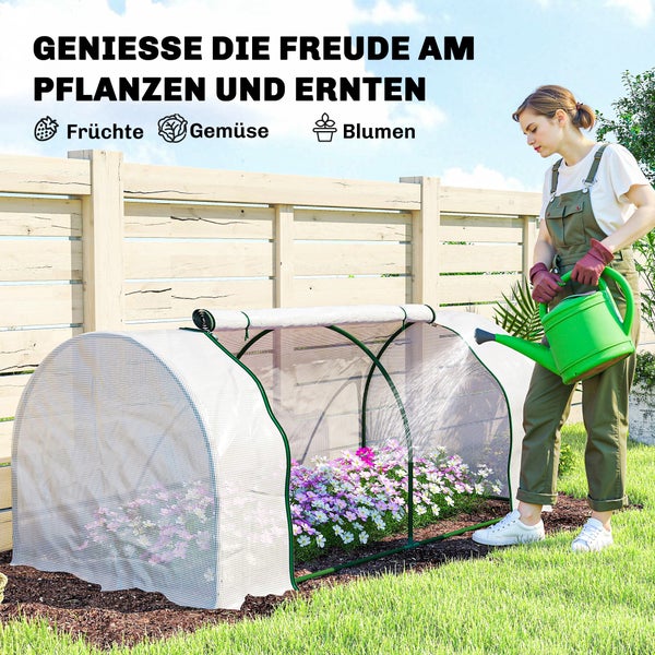 Eine Frau gießt Blumen in einem kleinen Foliengewächshaus im Garten vor einem Holzzaun.