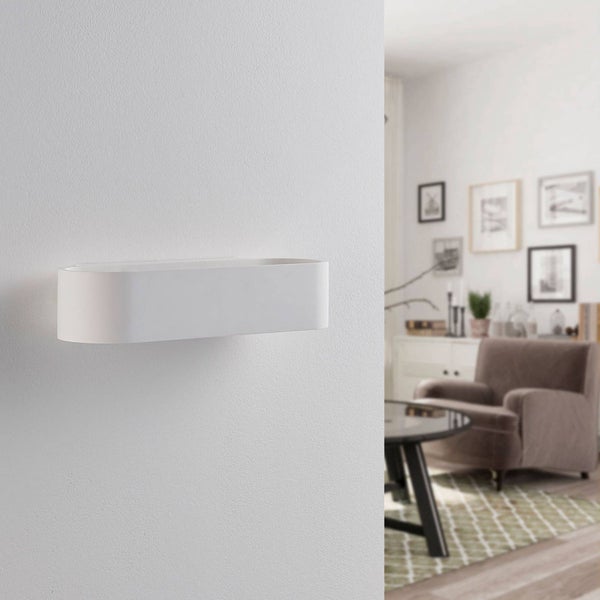 Ovale, weiße Wandleuchte an einer Wand montiert, im Hintergrund ein helles, modern eingerichtetes Wohnzimmer mit Sessel und Wandbildern.