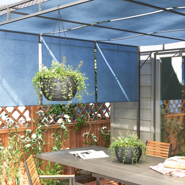Helle Szene mit blauer Pergola-Bespannung über einem Sitzbereich im Freien mit Gartentisch, Hängeampel und Blumen vor einem Holzzaun.