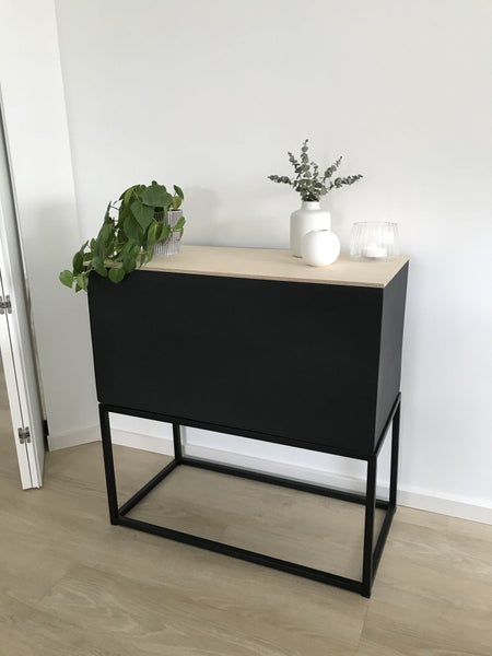 Modernes Sideboard mit schwarzem Korpus, heller Holzplatte und schwarzem Metallgestell, dekoriert mit Pflanze und Vasen in hellem Raum.