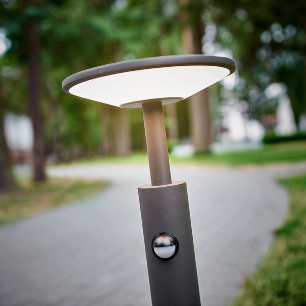 Moderne LED-Wegeleuchte aus Metall mit flachem Kopf und integriertem Bewegungsmelder an einem Gartenweg.