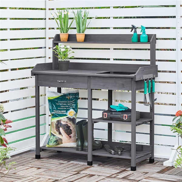 Gartentisch mit Ablageflächen, Schublade und Hakenleiste für Gartengeräte, dekoriert mit Pflanzen, Erde und Werkzeug.