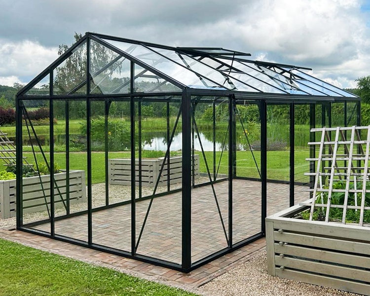 Modernes Gewächshaus mit schwarzem Aluminiumrahmen und Glaswänden in einem Garten mit Teich und Hochbeeten.