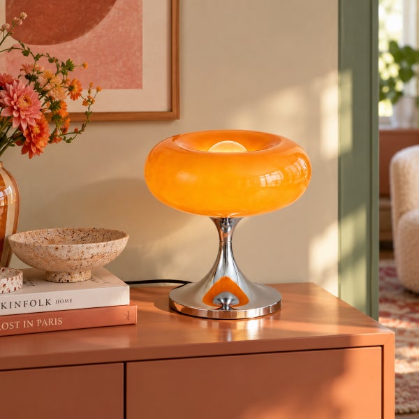 Moderne Tischleuchte im Retro Design mit orangefarbenem Glasschirm und glänzendem Metallsockel auf einer Kommode in einem hellen Wohnraum.