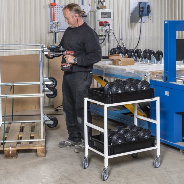 Ein Mann montiert Rollen an einem Wagen mit einem Akkuschrauber, während ein Rollwagen mit weiteren Rollen bereitsteht.