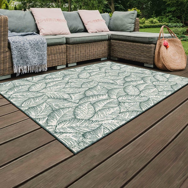 Rechteckiger Outdoor-Teppich mit weißem Blattmuster auf einer Holzterrasse neben einem Lounge-Sofa aus Geflecht.