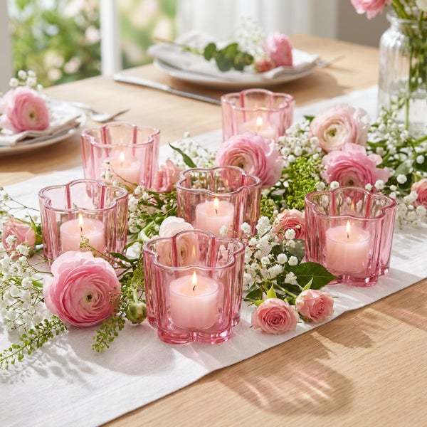 Festliche Tischdekoration mit mehreren Glaswindlichtern und frischen Blumenbouquets auf einem hellen Tischläufer.