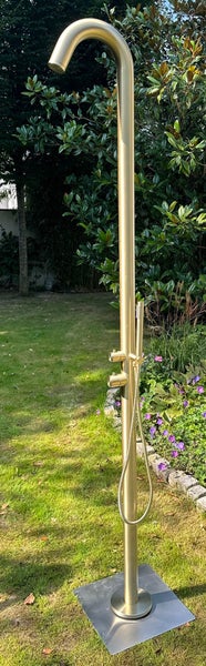 Freistehende goldfarbene Gartendusche aus gebürstetem Metall mit Handbrause und gebogenem Duschkopf in einem Garten.