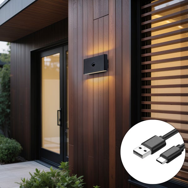 Moderne schwarze LED-Außenwandleuchte mit Bewegungsmelder an einer dunklen Holzfassade neben einer Glastür, inklusive USB-Ladekabel.