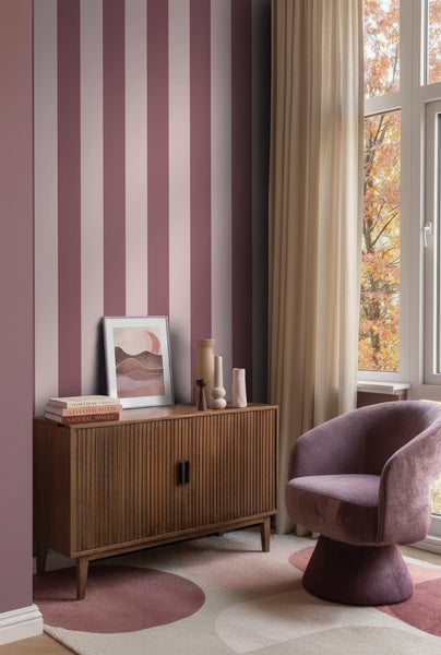 Vliestapete mit vertikalen Streifen hinter einem Sideboard aus Holz in einem hellen Wohnzimmer mit Sessel und Teppich.