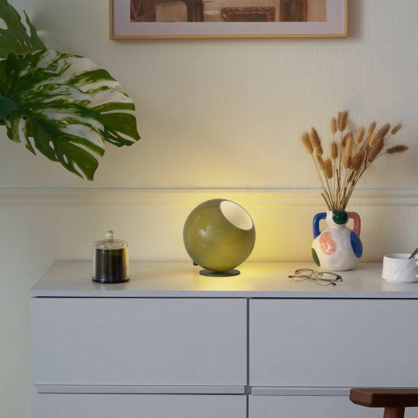 Moderne kugelförmige Tischleuchte in Olivgrün, leuchtend auf einer weißen Kommode neben einer Vase mit Trockenblumen und einer Brille.