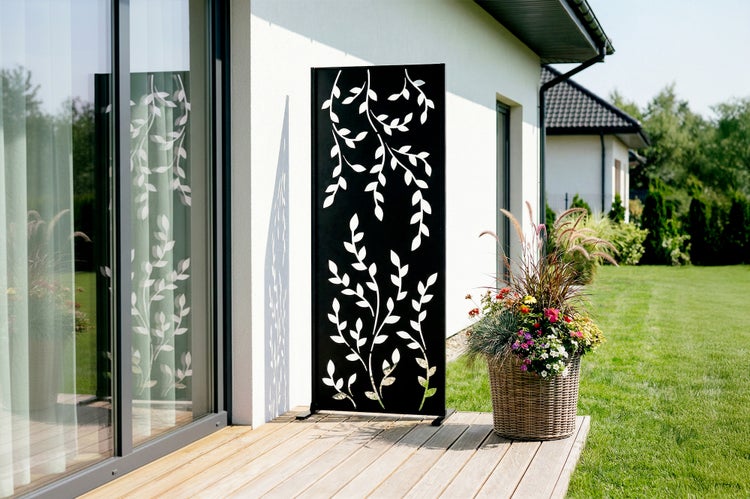 Dekorativer Sichtschutz aus Metall mit Blattmuster auf einer Holzterrasse neben einem Blumentopf und einer Hauswand.
