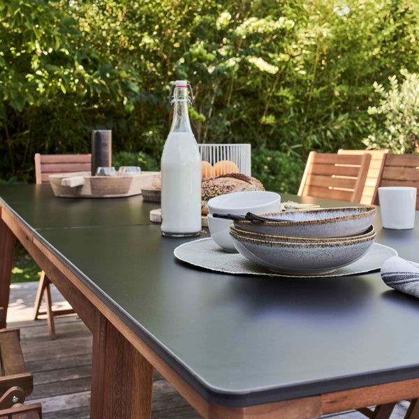 Gedeckter Gartentisch mit dunkler Tischplatte und Holzgestell auf einer Holzterrasse im Garten, umgeben von Gartenstühlen aus Holz.