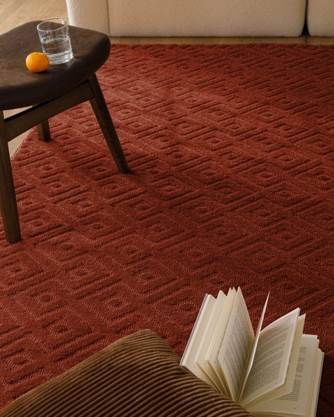 Strukturierter rostroter Teppich mit Rautenmuster in einem hellen Wohnzimmer mit Holzstuhl, Sofa und aufgeschlagenem Buch.