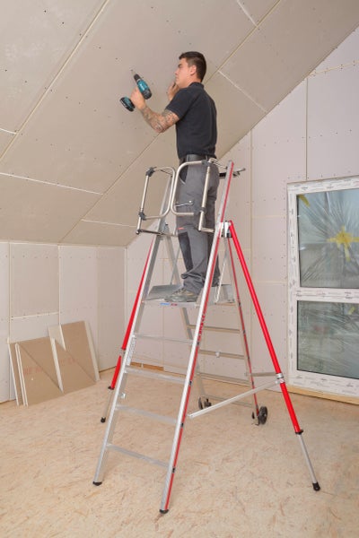 Handwerker auf einer Arbeitsbühnenleiter beim Anbringen einer Gipskartondecke mit einem Akkuschrauber in einem Raum.