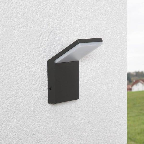 Moderne LED-Außenwandleuchte mit anthrazitfarbenem Metallgehäuse und geneigtem weißem Lichtdiffusor, montiert an einer strukturierten weißen Wand.