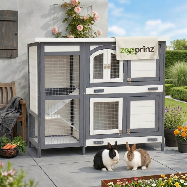 Großer zweistöckiger Kleintierstall aus Holz in Grau und Weiß mit Rampe, Gittertüren und Zooprinz Logo, auf einer Terrasse mit zwei Kaninchen.